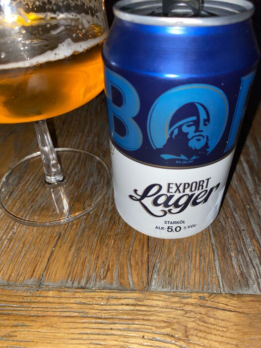 En ölburk och ett halvfullt ölglas på ett träbord. Burken visar en logotyp och texten "Export Lager".