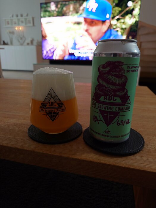 Ett glas öl framför burk, Apex Brewing logo, bokeh TV-bakgrund, inomhus hemmiljö.