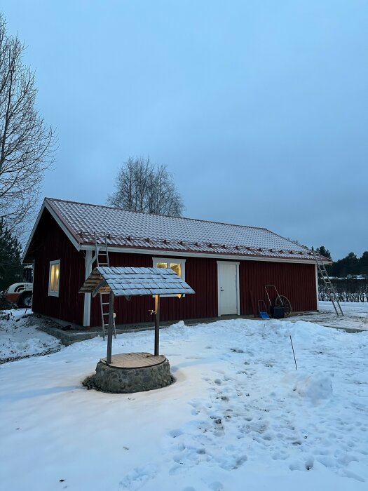 Röd stuga, snötäckt mark, brunn med tak, tidig vinterkväll, lugn atmosfär, träd bakgrund.