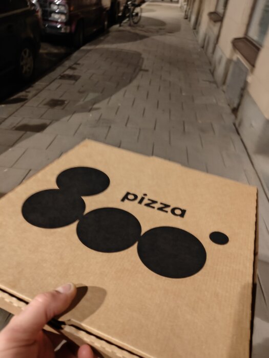 Person håller en pizzakartong på en stadsgata om natten. Grafik på lådan föreställer abstrakt design.