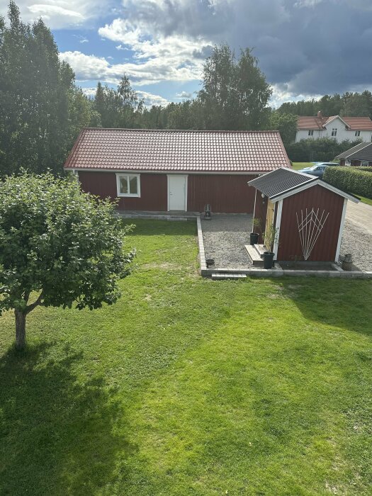 Rött lantligt hus med tegeltak, grönt gräs, trädgårdsväg och växande träd, molnig himmel.