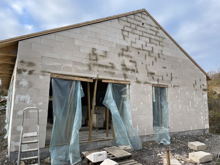 Byggnad under konstruktion med grå betongblock, stege, och blåa plastskydd över dörröppningen.