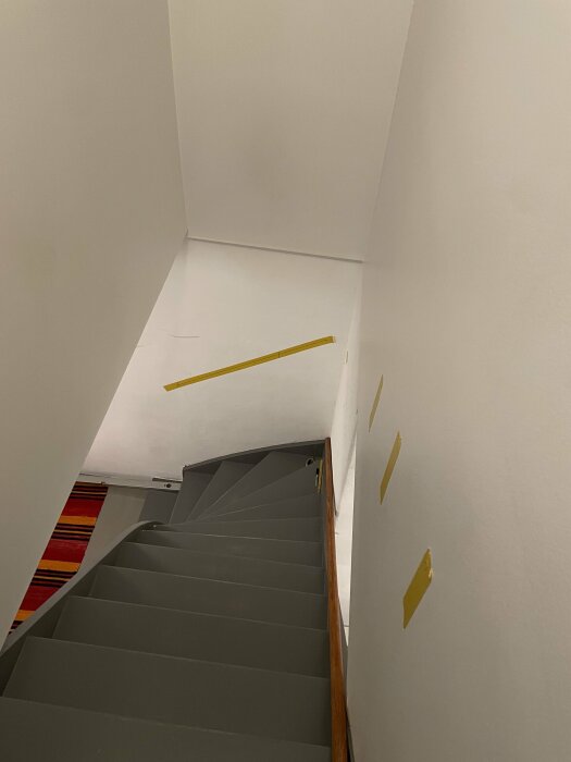 En trappa med grå steg, träledstång, vita väggar och gula linjer på väggen.