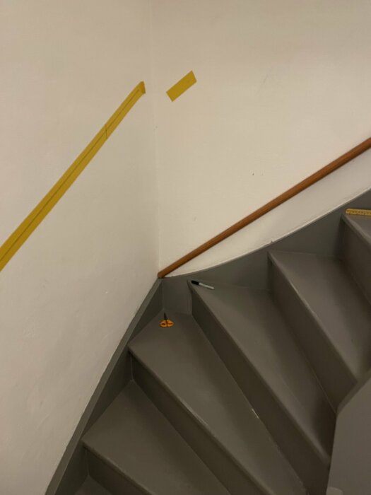 Trappa med gråa steg, gul räckesskena, vit vägg, lösa gula markeringstejper och bruna handledare.