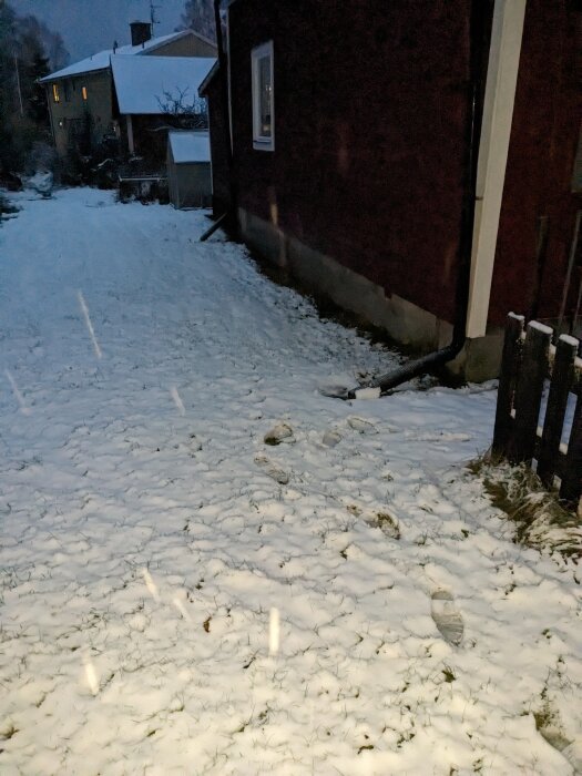 Snötäckt mark med fotavtryck, röd husvägg, fönster, tidig kväll eller morgon.