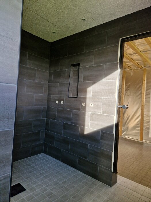 Ett ombyggt rum med kaklade väggar och golv, ett duschområde under konstruktion, utan armaturer installerade ännu.
