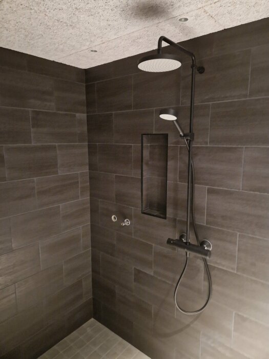 Modernt badrumsduschutrymme med mörkt kakel, stort duschhuvud och handhållen dusch i svart.