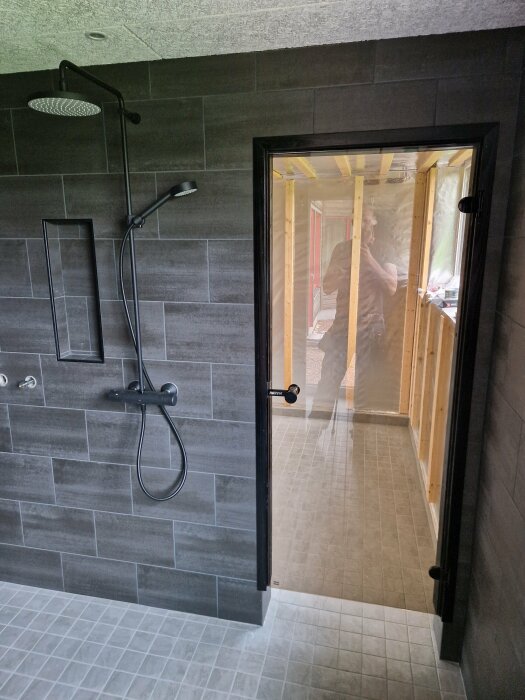 Modernt badrum med grå kakel, dusch, glasdörr och suddig figur i nästa rum.