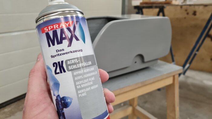 Hand håller en burk Spray Max 2K grundfärg framför en icke helt monterad bilkofångare på arbetsbänk.