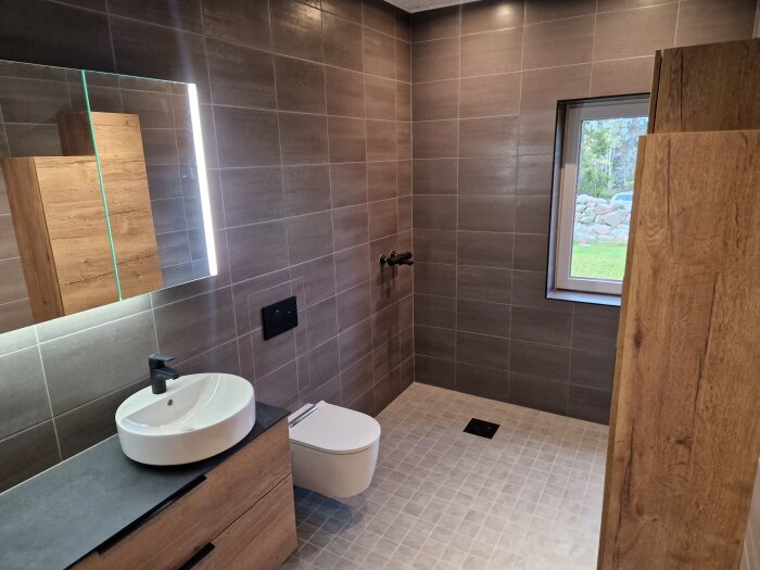 Modernt badrum, trä och kakel, spegel, handfat, toalett, fönster med utsikt, diskret belysning.