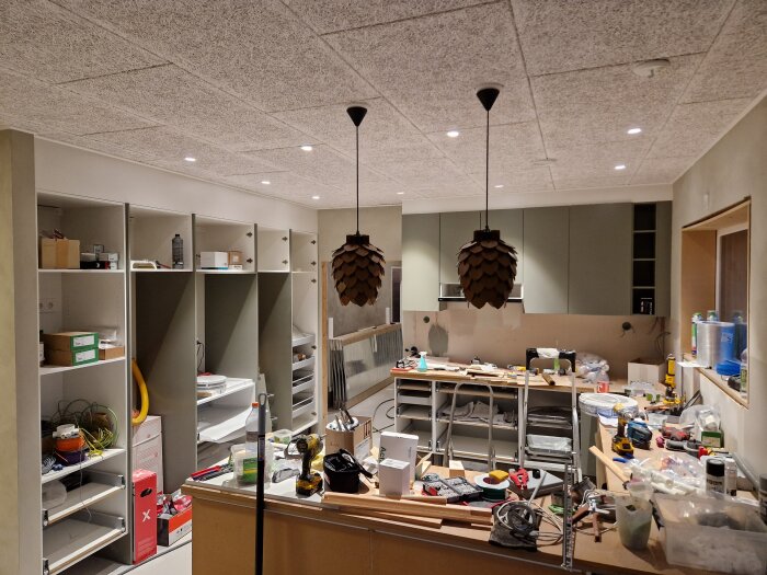 Oorganiserat rum under renovering med verktyg, material, hyllor och hängande lampor.
