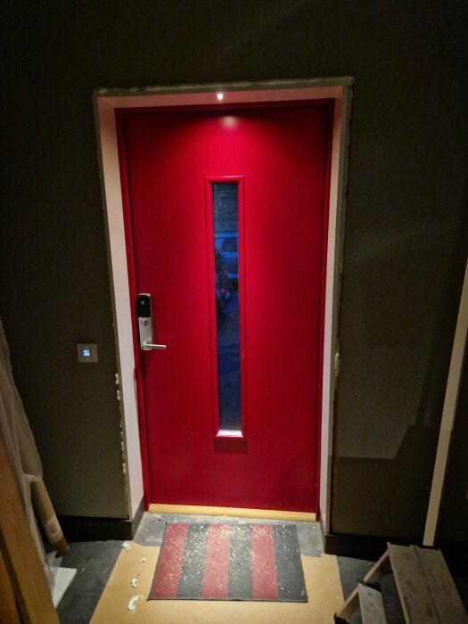 Röd dörr med smalt fönster, elektroniskt lås, omgiven av mörka väggar, till synes inne i byggnad.