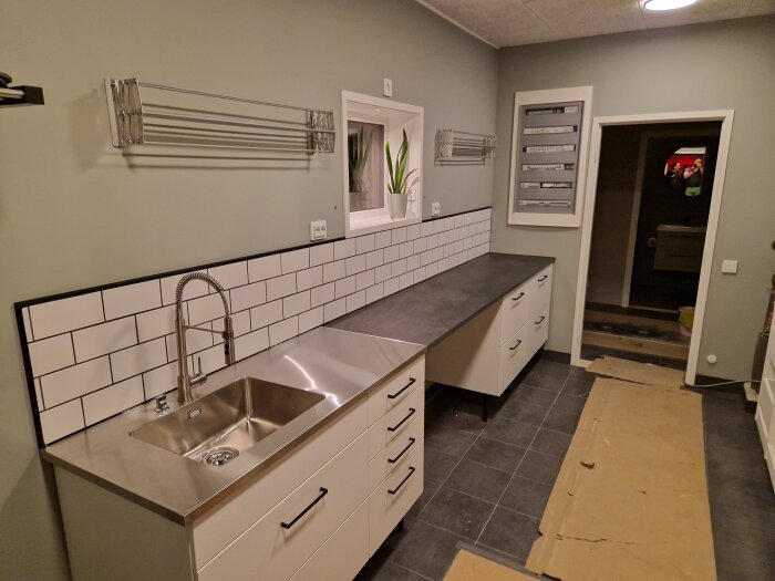 Modernt kök under renovering med vita skåp, rostfri diskho, och kakelvägg.