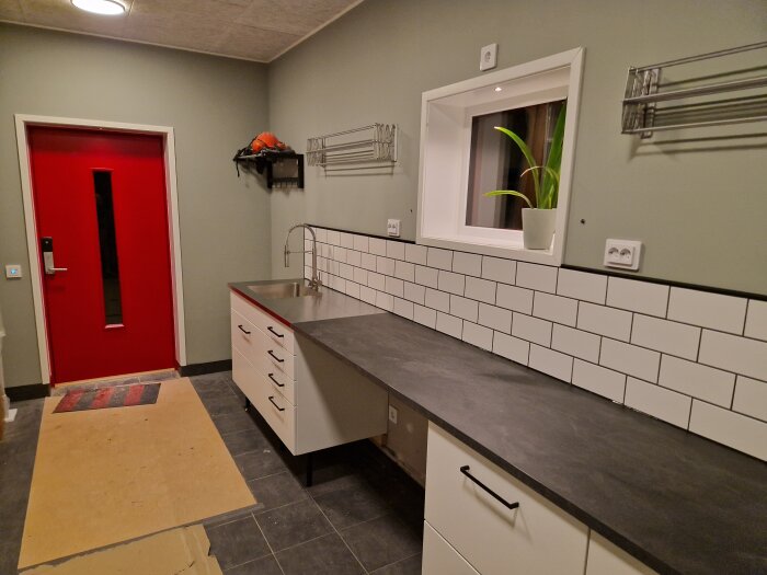 Ett modernt kök under renovering med vita skåp, svart bänkskiva, röd dörr och gråa väggar.