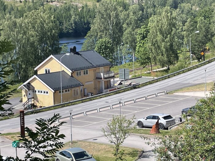 Soligt bostadsområde med gult hus, parkering, bilar, träd, flod i bakgrunden.