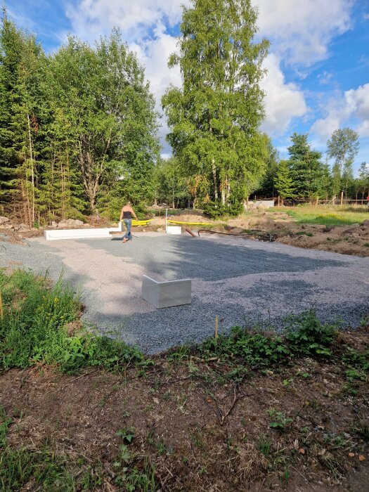 Byggplats i skogsglänta, grusgrund, person bär virke, blå himmel med moln, gröna träd.