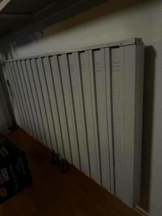 En stor vit radiator upphängd på vägg i rum med trägolv, med ljus tapet och kartong på golvet.