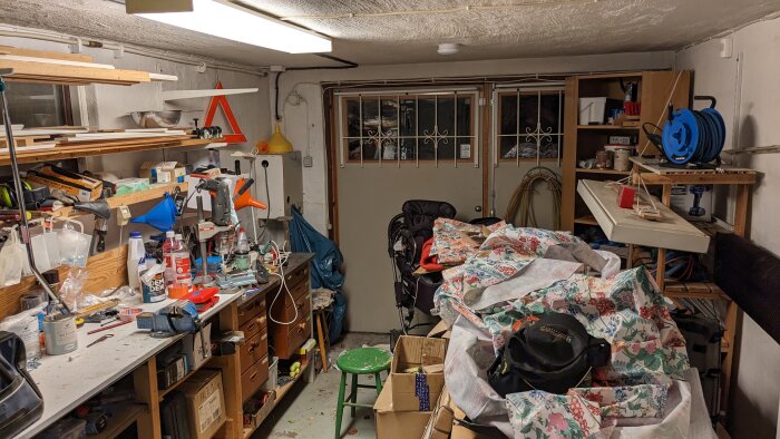 Oorganiserad källarverkstad med verktyg, trähögar, barnvagn och oreda på arbetsbänkar och hyllor.