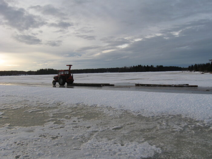Röd traktor på frusen sjö, snötäckt is, träbryggor, skymningshimmel, skog i horisonten.