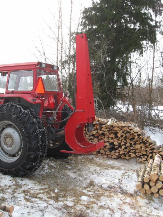 Röd traktor med vedklyv, snöig mark, staplad ved, träd i bakgrunden.