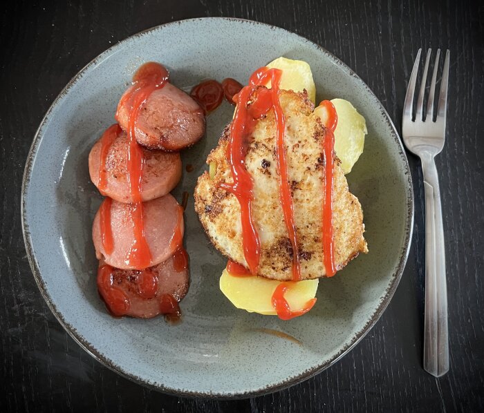 Stekt kycklingfilé och korvskivor med ketchup på tallrik, mörk bakgrund, gaffel på sidan.