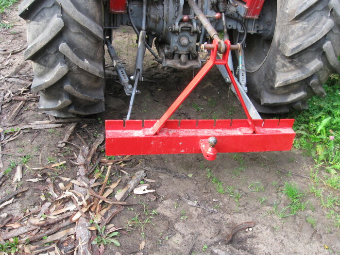 Röd jordfräs kopplad till traktor, står på jordbruksmark med gräs och kvistar.