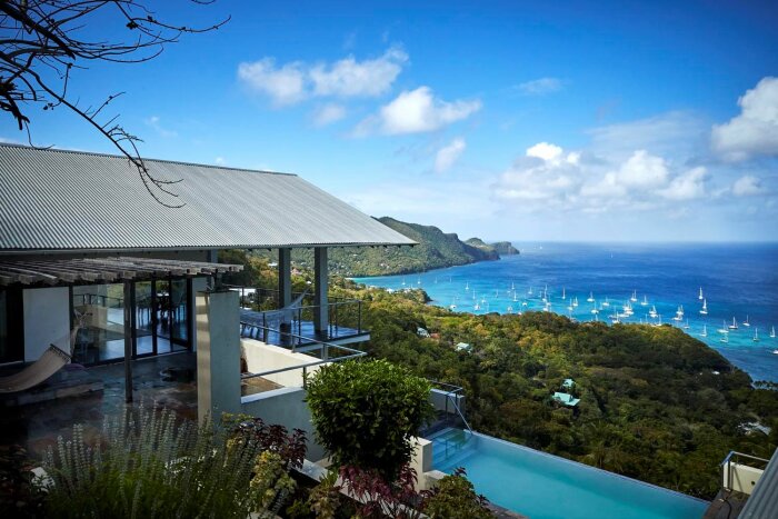 Modernt hus med terrass, pool, utsikt över hav och segelbåtar, tropisk miljö.
