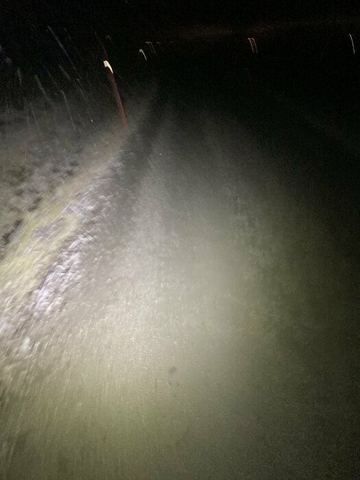 Mörk nattväg belyst av fordonsstrålkastare, regn eller snöflingor i ljuset, suddig och mörk omgivning.