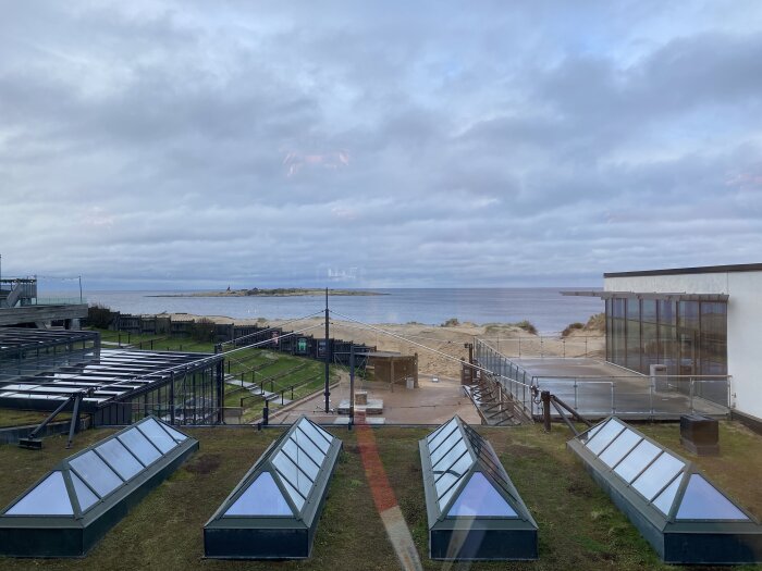 Kustlandskap, molnig himmel, strand, havsutsikt, takfönster, terrass, gräsmatta, spegling av inomhusbelysning, inga människor.