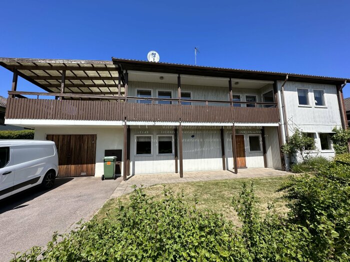 Tvåvåningshus med balkong, garage, vit skåpbil, klar himmel och grönt gräs.