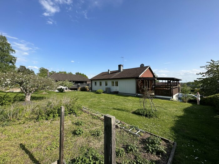 Ett hus med veranda, grönskande trädgård och klarblå himmel. En person synlig till höger.