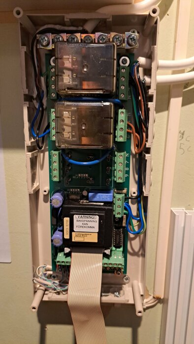 Elektriskt kopplingsskåp med reläer, varningsetikett, kablar och terminaler, öppet på vägg.
