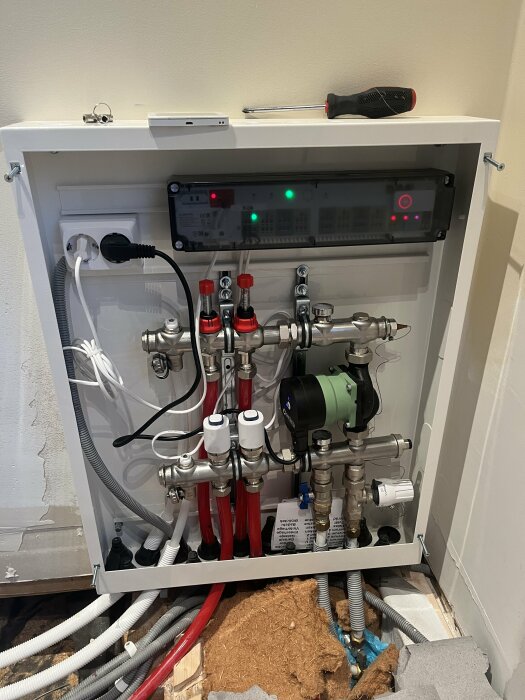 Ett öppet uppvärmningssystemskåp med rör, ventiler, pump och elektronik. Skruvmejsel och mobilladdare ovanpå.