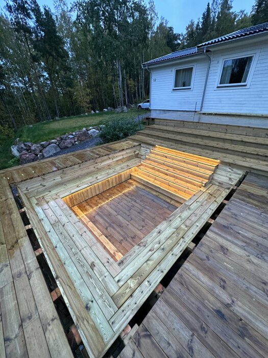 Trädäck under konstruktion, belysning installerad, hus och träd i bakgrund, kväll, vått trädäck, grå himmel.