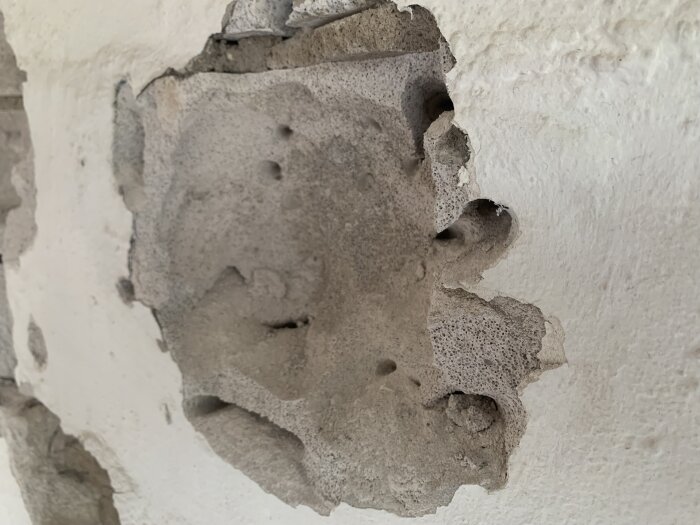 Förvitrad vägg med skador, avflagning. Texturer och hål synliga. Närbild på skadad betong eller puts.