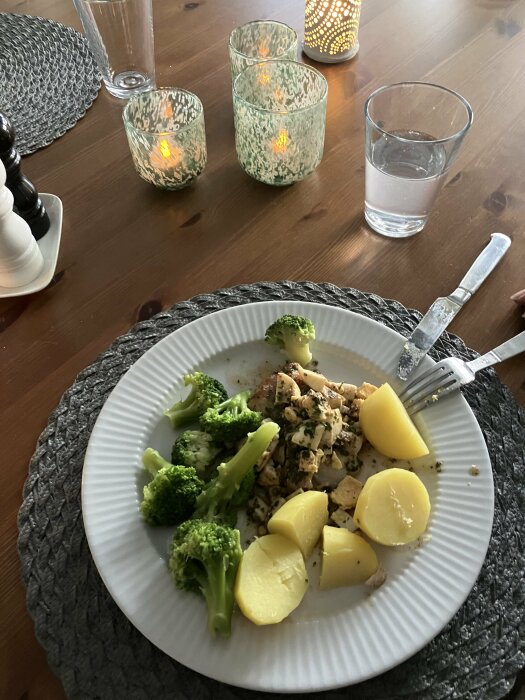 Måltid med kyckling, potatis, broccoli. Ljuslyktor tända. Vattenglas. Middagsbord. Schackpjäs synlig. Mörk träyta.