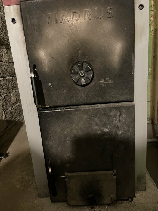 En smutsig, svart gjutjärnskamin märkt "VIADRUS" i ett industriellt eller källarrum.
