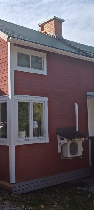 Rött hus, vita fönsterkarmar, luftkonditionering, tegelskorsten, grönt gräs, delvis molnig himmel.