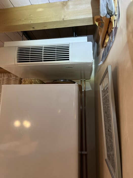 Luftkonditioneringsenhet ovanför kylskåp, träbjälkar, spegelreflexion och vägg med bilder, inomhus.