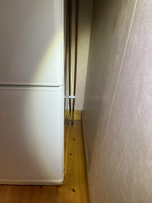 Vit kylskåp, trägolv, kablar längs väggen, hörn av ett rum, dagsljus.
