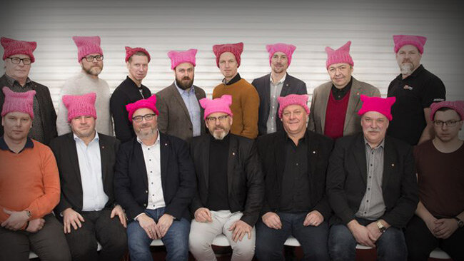 Grupp män i olika åldrar bär rosa stickade mössor med kattöron, poserar för fotografi, neutral bakgrund.
