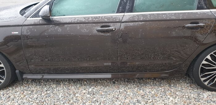 Sidosyn av brun bil täckt med vattendroppar på stenbelagd mark. Fahrverke.Hjulen synliga. Speglar. Inga personer.