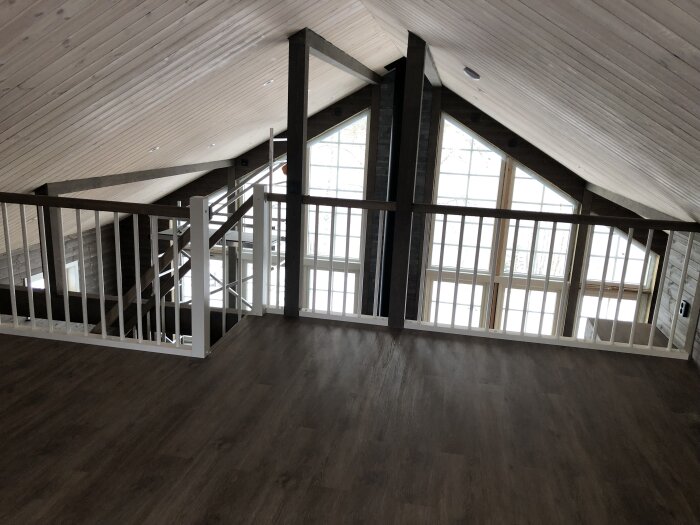 Inomhus vy av ett loft med träbjälkar, vit panel, mörkt golv, och stora fönster.