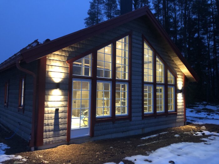 Trähus med tända fönster i kvällsljus, snöfläckar på marken, skog i bakgrunden.