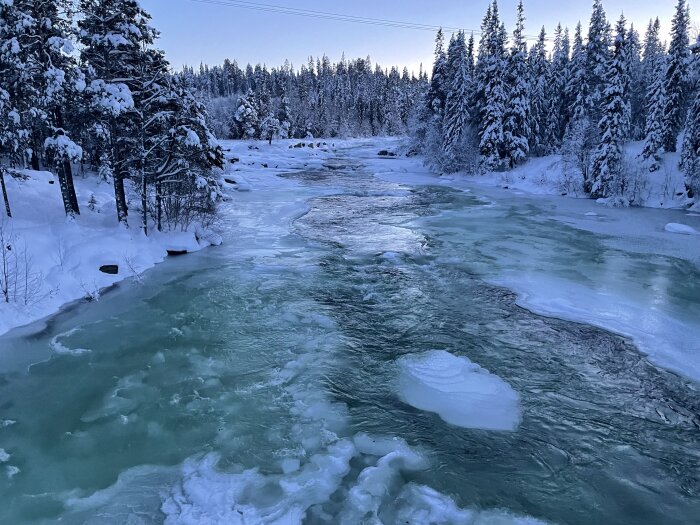 Vinterlandskap med fruset flod, isformationer, snötäckta träd och blå himmel.