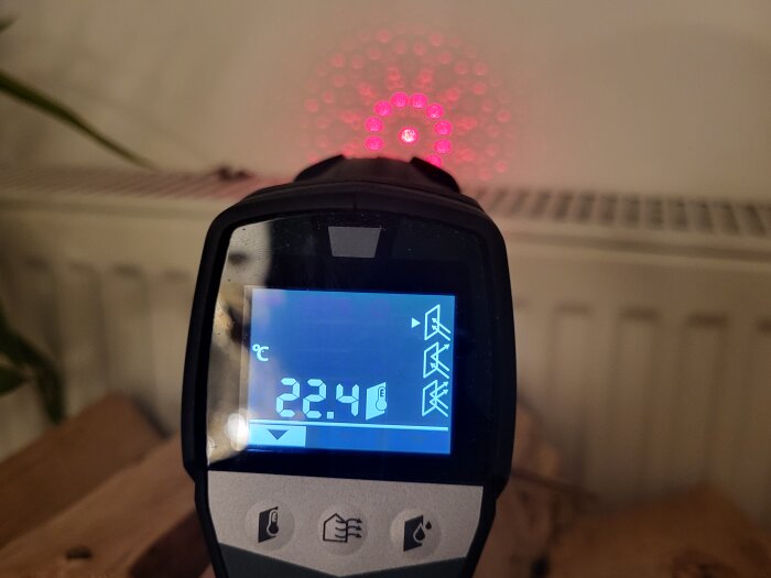 Infraröd termometer visar 22.4°C mot en bakgrund av rött laserstrålemönster och suddig inredning.