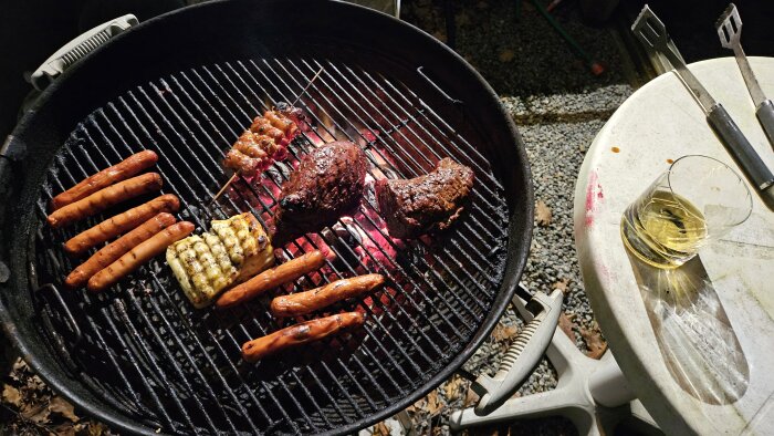 Grill med kött, korvar, majs; grilltång; ett glas på bordskant nattetid.