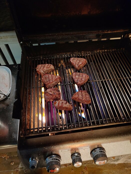 Köttbitar som grillas på en gasolgrill med synliga lågor och reglage i förgrunden.