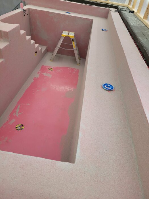 Byggarbetsplats med rosa isolering och stege i tom swimmingpool under konstruktion.