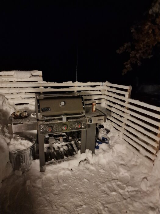 Gasolgrill täckt med snö på en terrass om natten, omgiven av en snöig miljö.